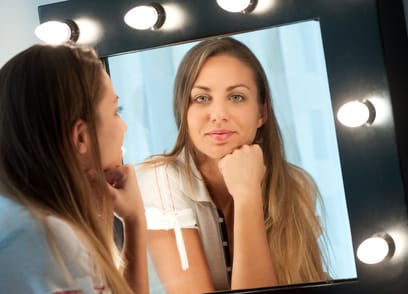 Attractive young girl with long brown hair sitting at a dressing table staring into the mirror with a thoughtful expression, focus to her reflection